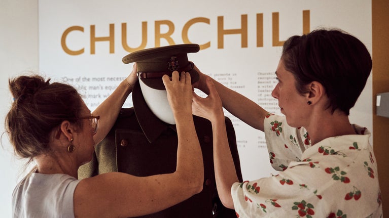 Two conservators adjust a hat at on a Winston Churchill mannequin beneath lettering on the wall which says Churchill.